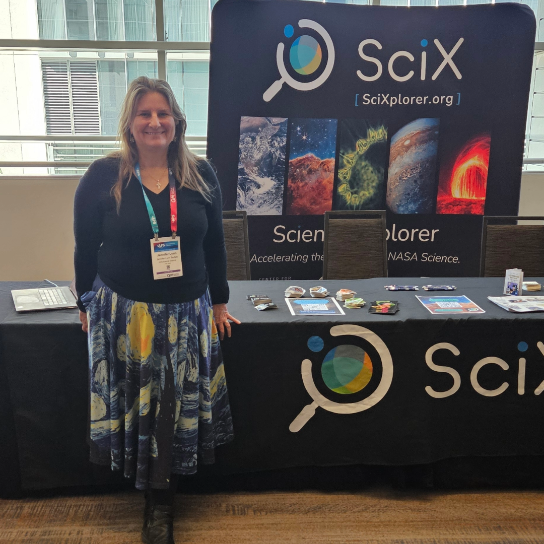 A woman stands smiling in front of a SciX / SciXplorer conference booth inside a bright building. She is wearing a dark sweater, a colorful galaxy-patterned skirt, black tights, and boots, with a conference badge hanging on a lanyard. The booth table behind her is covered with a black tablecloth featuring the SciX logo and images of space and planetary phenomena, along with brochures and small promotional items spread across the table.